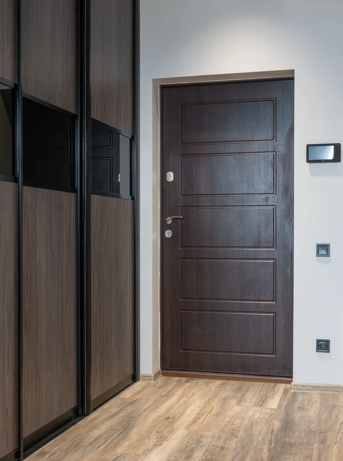 A short entry hall with a weathered oak-look laminate floor running past dark wood-grain sliding wardrobe doors to a dark paneled front door
