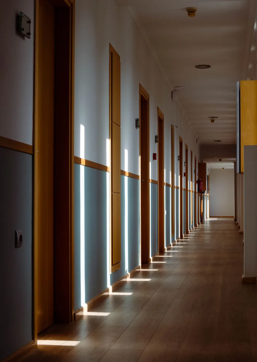 A long corridor lined with wooden doors and blue-grey lower walls, with light wood-look flooring and afternoon sun falling across it