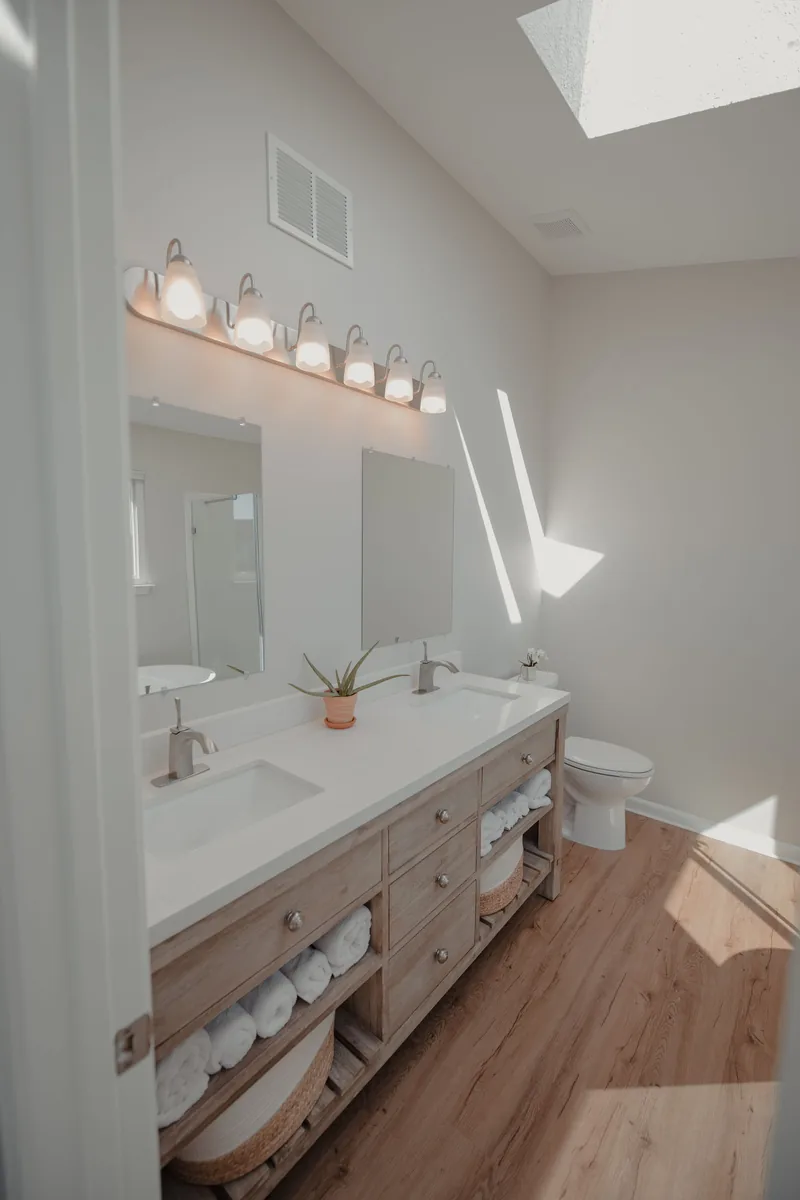 A bright bathroom with a double vanity, a toilet and light wood-look plank flooring lit by a skylight