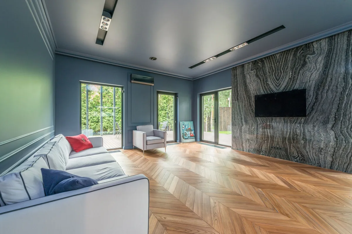 A living room with light wood-look flooring laid in a chevron pattern, a grey sofa, and glass doors opening onto a patio and garden