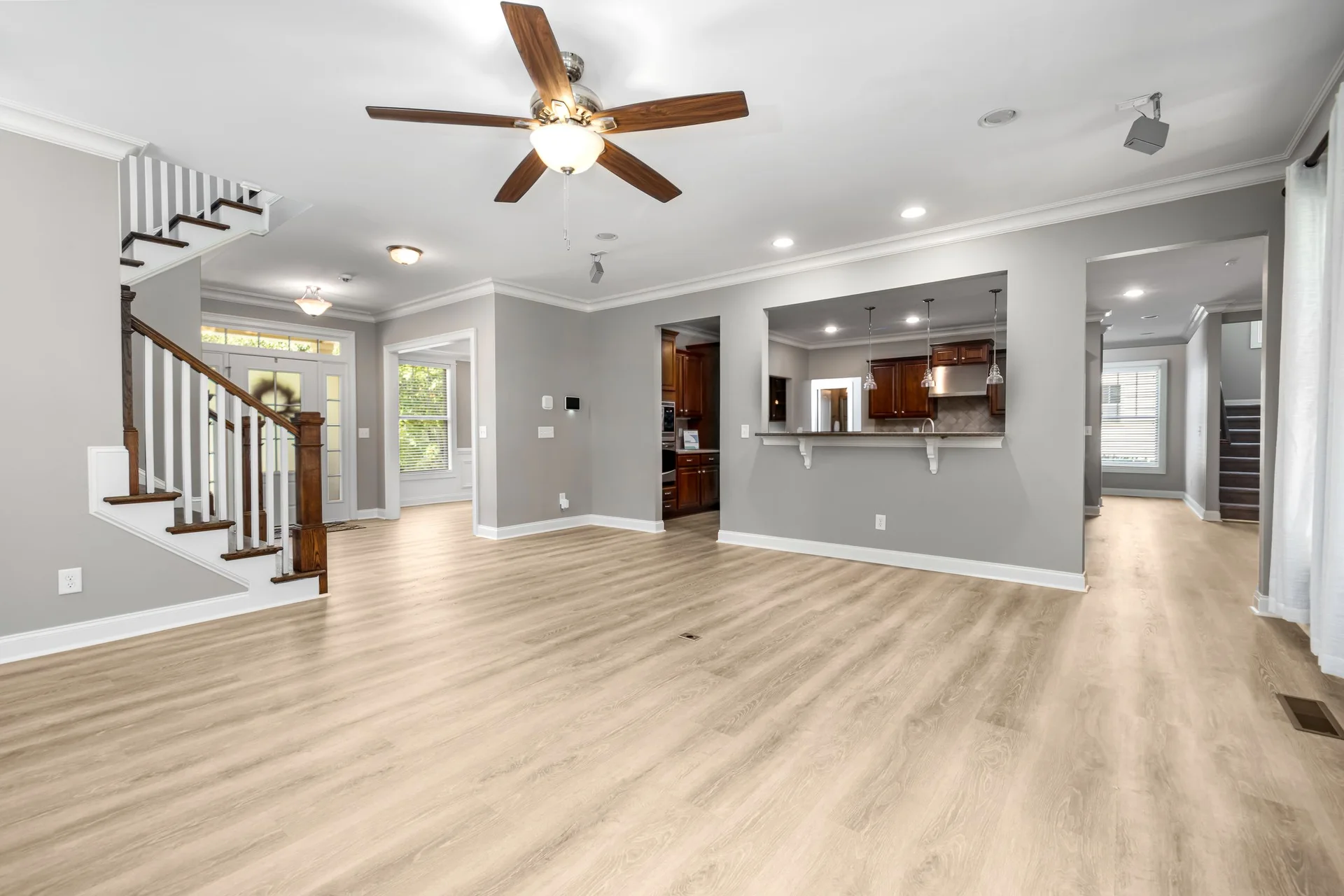 An empty open-plan living room with light oak-look vinyl plank running unbroken from the front entry and staircase through to the kitchen pass-through and a back hallway
