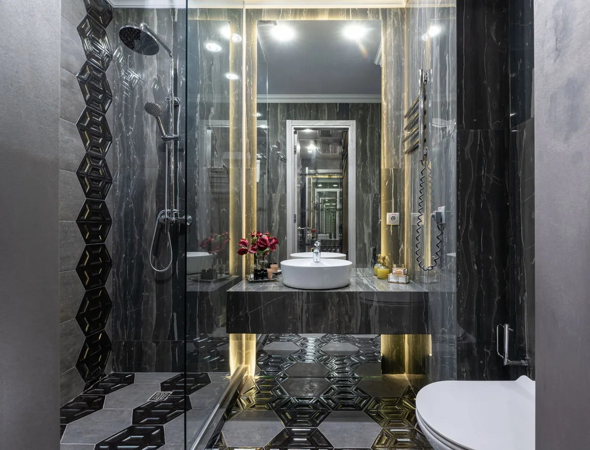 A narrow modern bathroom with dark marble-look wall tile, a frameless glass walk-in shower, hexagon floor tile and a vessel sink under a backlit mirror