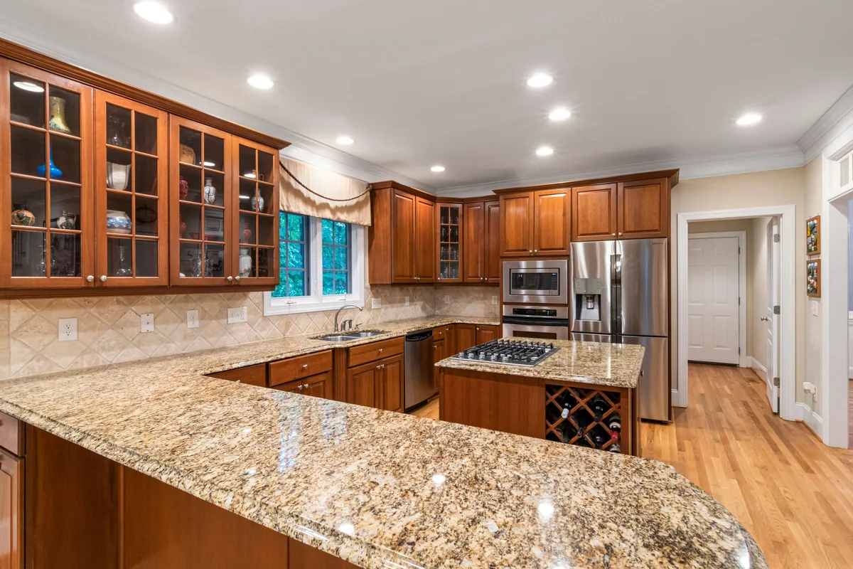 A large kitchen with speckled tan granite countertops on a peninsula and island, cherry wood cabinets and a light hardwood floor